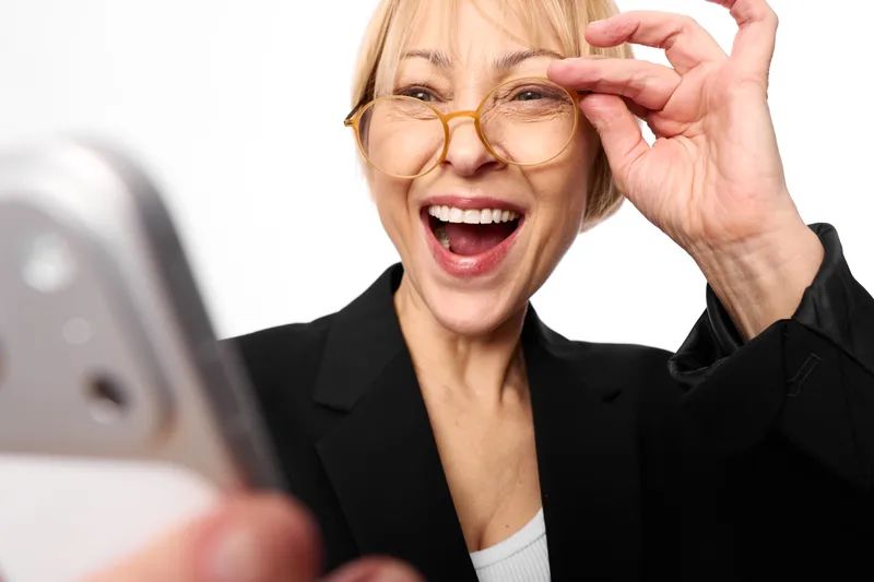 Cheerful woman in office attire laughing, adjusting glasses with her hand, blurred laptop foreground, bright workspace, joy and excitement, communication with technology