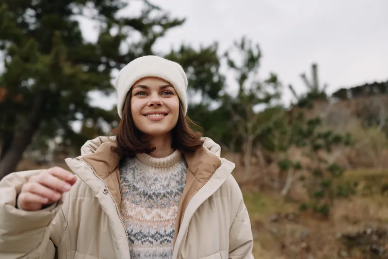 Woman in a warm jacket and beanie smiles outdoors with a natural landscape behind her, capturing a cozy, candid moment in cool weather and enjoying fresh air.