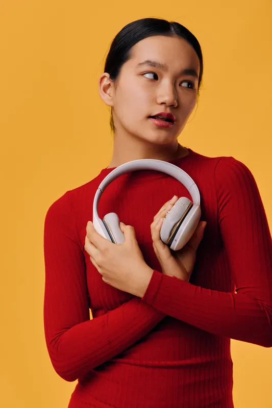Young woman holding white wireless headphones in hands against yellow background wearing red sweater. Portrait of female with dark hair looking sideways in studio setting.