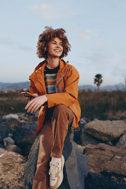 Smiling woman with curly hair wearing an orange jacket, relaxing on rocky ground outdoors. Scenic landscape and nature background create a calm lifestyle mood in soft sunset light.