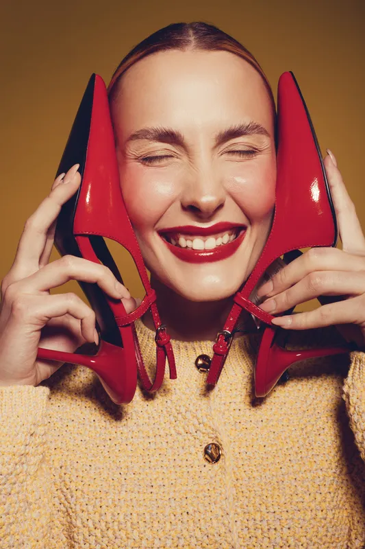 Smiling woman with closed eyes holds red high heel shoes near face, wearing yellow knitted sweater. Concept of fashion, happiness, style, and joyful emotions in studio lighting.
