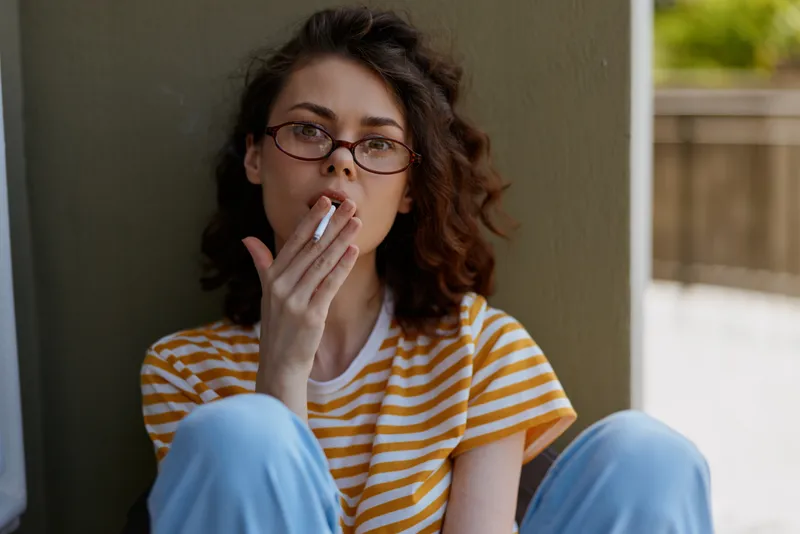 Woman smoking, casual lifestyle in a homey setting, portrait with glasses, striped shirt, relaxed mood, candid moment of everyday life, warm daylight and thoughtful expression.