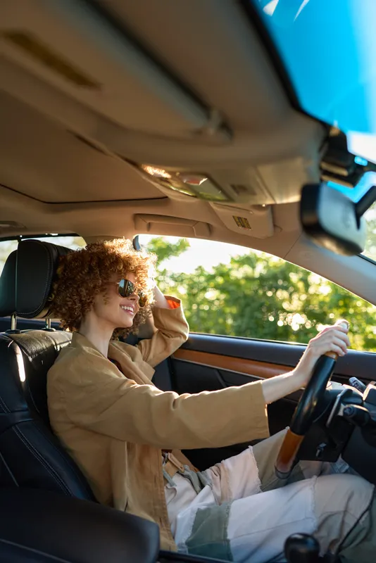 Cheerful woman driving a car, sunglasses on, warm sunlight, casual jacket, candid travel moment, retro film color mood, relaxed pose, vibrant scenery