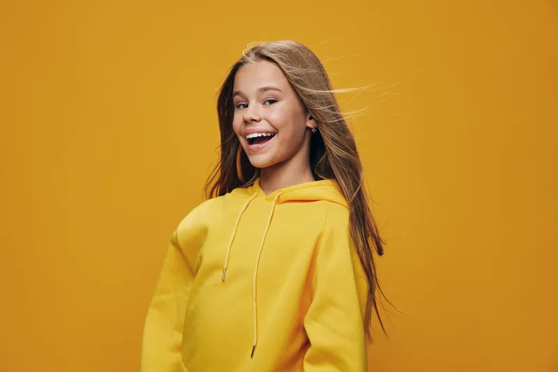 Girl in yellow hoodie smiling and posing against orange background with long hair flowing, cheerful expression on face for happy youth and casual fashion concept.