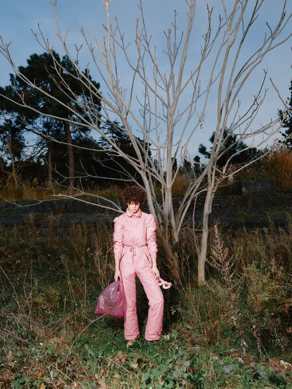 Pink jumpsuit worn by a model stands in a field with bare trees, autumn scenery and a pink bag, creating bold color contrast and urban fashion photography vibe.