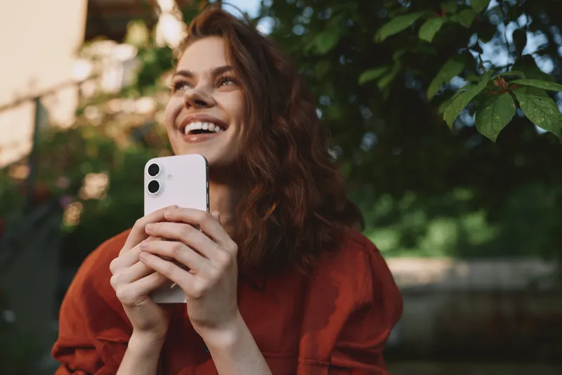 A cheerful woman in a red top smiles while holding a smartphone outdoors, capturing a candid lifestyle moment and joyful everyday connection