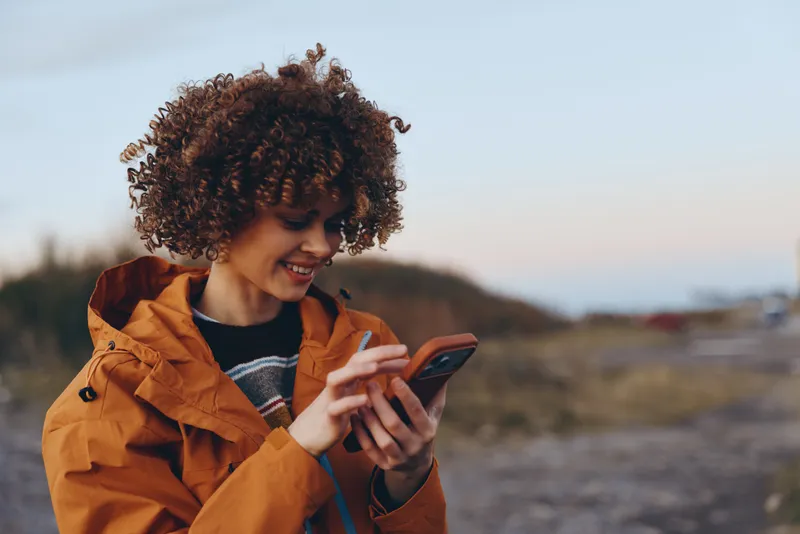 Woman lifestyle portrait outdoors, curly hair, orange jacket, smiling, holding smartphone, reading screen, soft blur background, open air walk near field at sunset