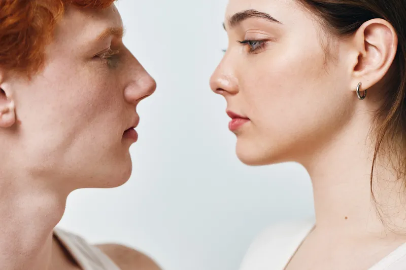 Portrait of young woman and man facing each other with neutral expressions on light background closeup. People, profile, skin and emotion with intimacy and soft natural lighting.