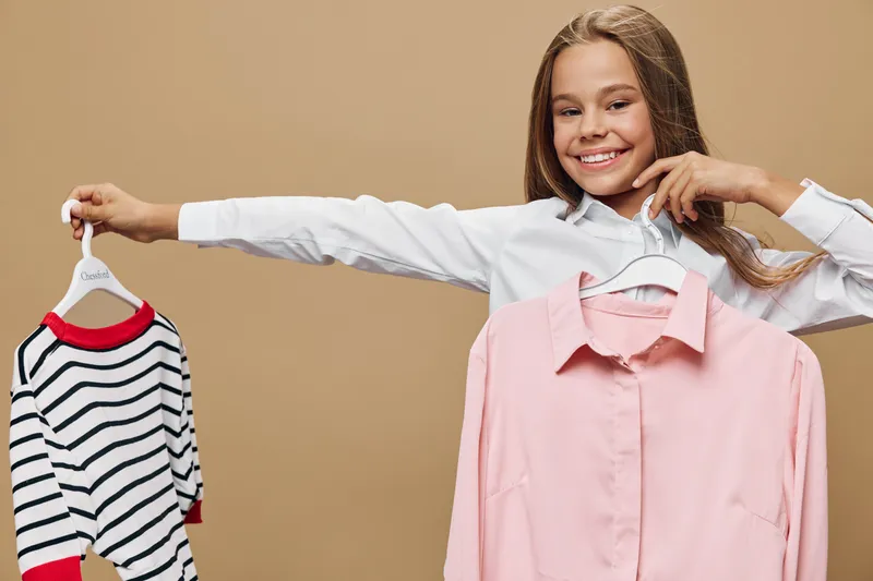 Girl smiling and holding two hangers with clothes including pink shirt and striped sweater for fashion and choice in studio with brown background