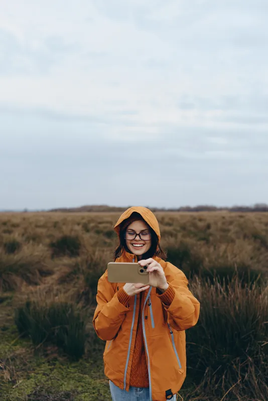 Smiling woman outdoors in a field, orange jacket and glasses, holding a smartphone. Casual lifestyle moment looking at the screen, natural daylight and relaxed travel mood in rural area