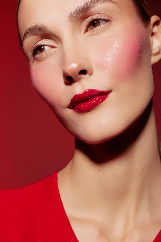 Close-up of a woman with red lipstick and blush on her cheeks wearing a red top against a red background. Concept of beauty, makeup, fashion, and skincare.