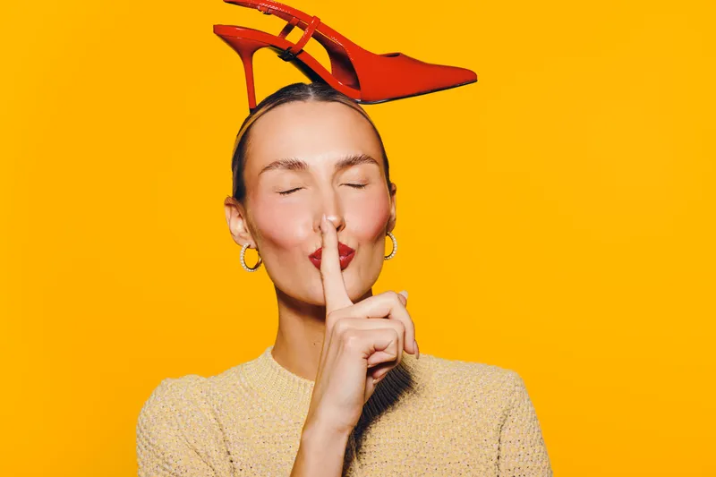 Woman with red high heel shoes on head makes silence gesture with finger on lips. Bright yellow background and casual beige sweater create playful and stylish mood.