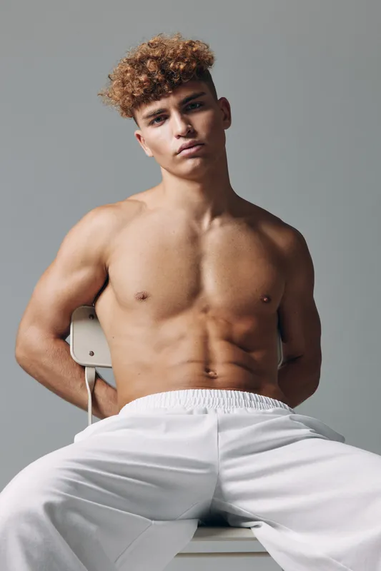 Young muscular man with curly hair sitting shirtless on chair wearing white pants against gray background. Fitness, bodybuilder, confident and casual pose in studio lighting.