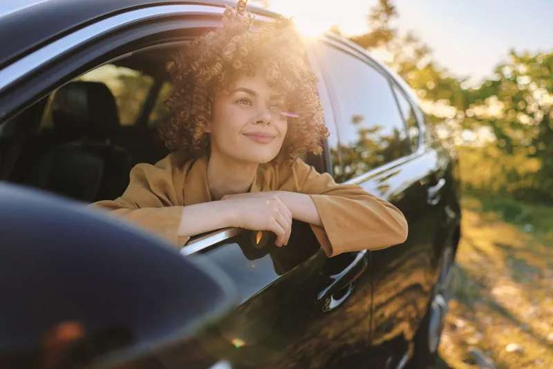 Cheerful woman leaning out of a car window, enjoying golden sunshine, carefree lifestyle moment, soft breeze, natural colors, vintage mood, outdoors ambient hues gentle sunlit landscape scene outdoor