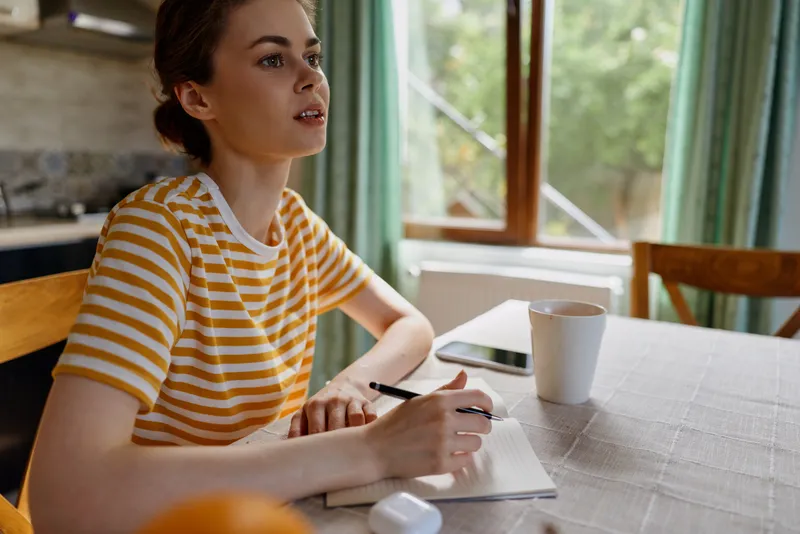 Woman lifestyle home writing notebook sunlight window casual interior cozy. A smiling woman sits at a table and writes ideas in a notebook, creating a peaceful daily routine.
