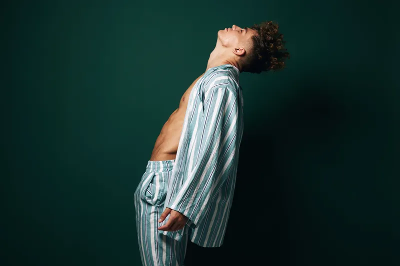 Man in striped pajamas leaning backward with eyes closed on dark green background. Relaxed young male model with curly hair in casual sleepwear for comfort and rest at home.