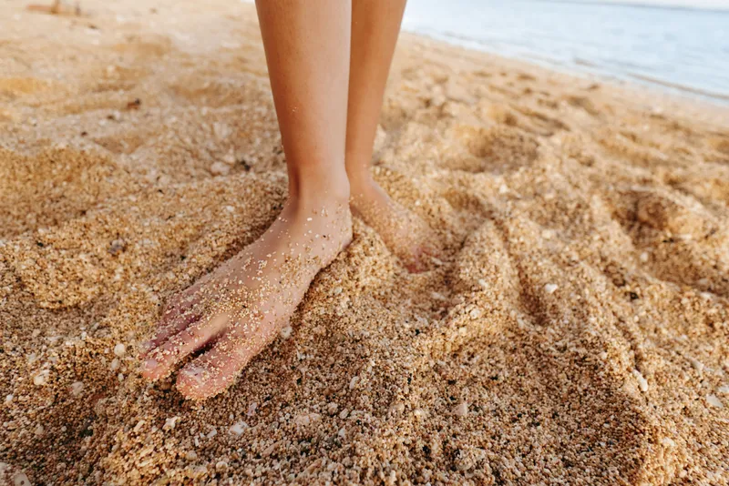 Close up of bare feet standing on sandy beach, warm sunlight, natural texture of sand grains, relaxed summer atmosphere, calm sea in background