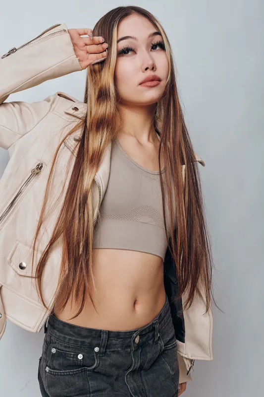 Young woman with long hair posing in fashionable beige jacket and crop top, showing confident expression and casual style against light background in studio.