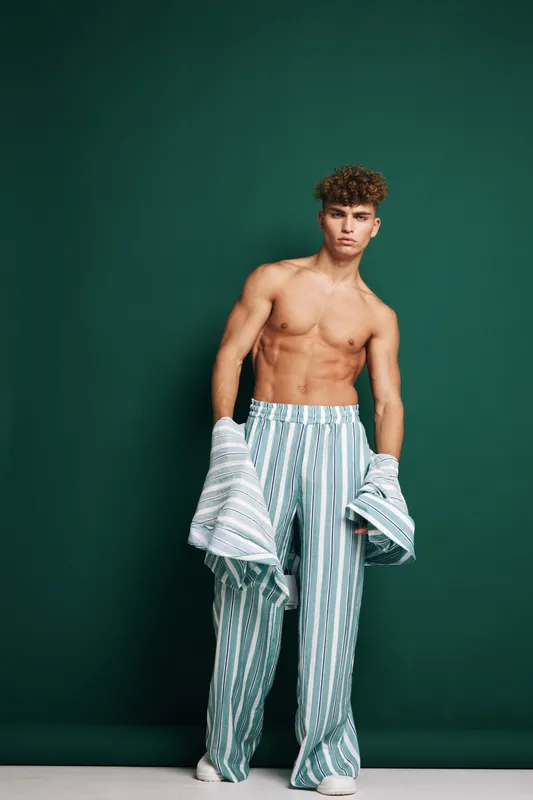 Young muscular man with curly hair posing shirtless in striped wide pants holding matching jacket against green background. Fashion model with confident expression and casual style.