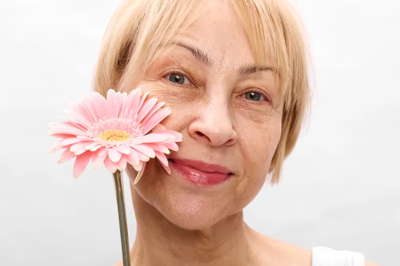 Elderly woman portrait, pink flower closeup, studio smile, blonde hair, holding the flower near cheek, soft light, white background, gentle look, calm atmosphere and natural skin tone