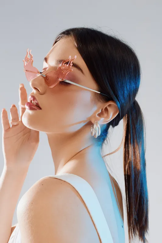 Young woman with sleek dark hair wears unique butterfly-shaped sunglasses and hoop earrings. She poses confidently in a white top against a neutral background. Concept for beauty fashion.
