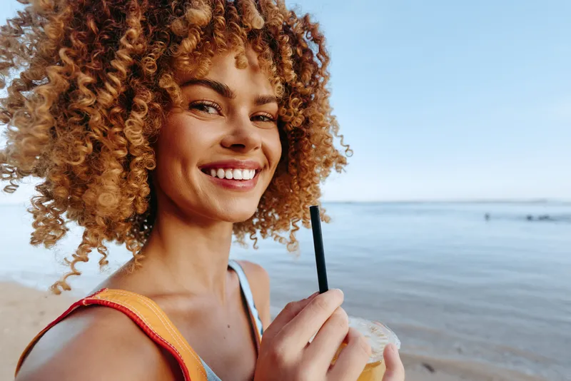 Young woman with curly hair smiles happily while holding a drink on a sunny beach with calm water and clear blue sky in the background.