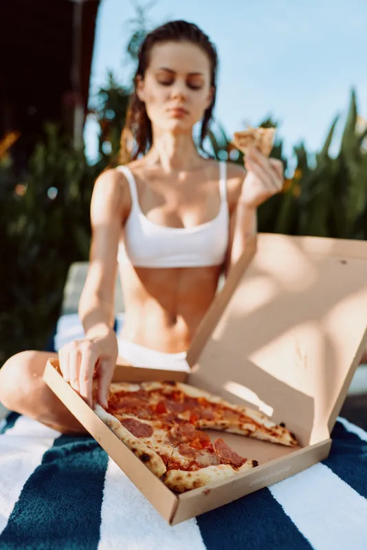 A young woman in a white crop top enjoys pizza outdoors on a sunny day, seated on a blanket with a box of slices nearby. A casual lifestyle moment of summer snacking and relaxation.