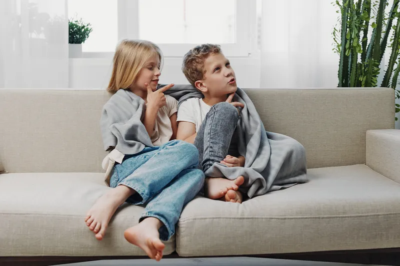 Two children wrapped in a blanket sit barefoot on a modern sofa, looking engaged and cozy in a bright living room with natural plants in the background.