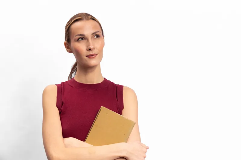 Thoughtful woman in sleeveless red top holds notebook, looks away on white background. Concept of planning, education, and focus on personal goals and productivity.