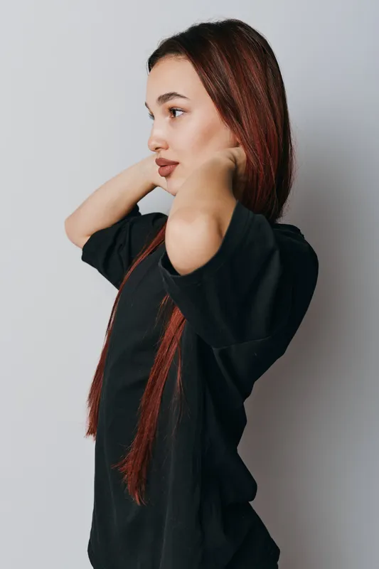 Young woman with long red hair wearing black shirt standing in profile against white background adjusting hair with hands behind neck. Casual style and natural look with focused expression.