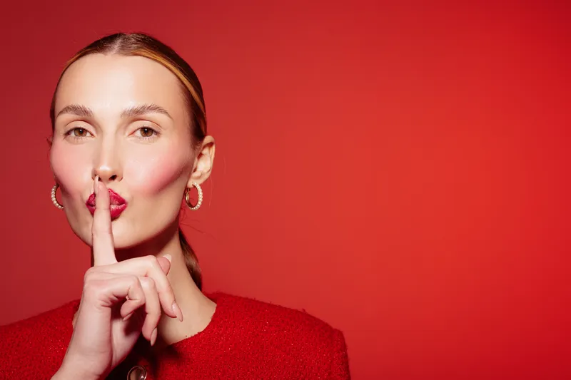 Portrait of a woman with red lipstick and earrings making a shushing gesture on red background. Concept for silence, secrecy, and quiet communication.