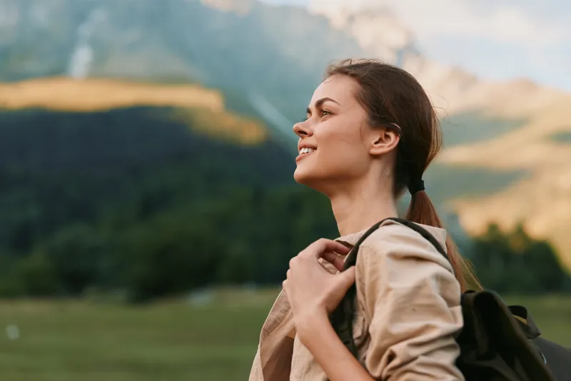 Woman traveler smiles while wearing a backpack in a meadow, with distant mountains, embodying outdoor lifestyle and exploration on a bright calm day. Scenic backdrop boosts mood.