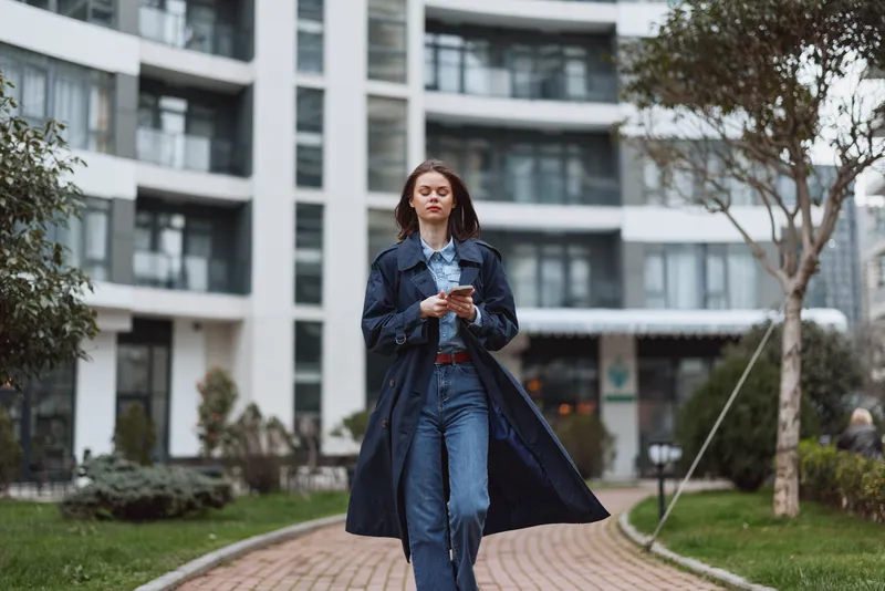 Young woman in denim jeans and trench coat walks outdoors, checking her phone as a light breeze moves her coat along a modern urban campus with glass walls reflecting light.