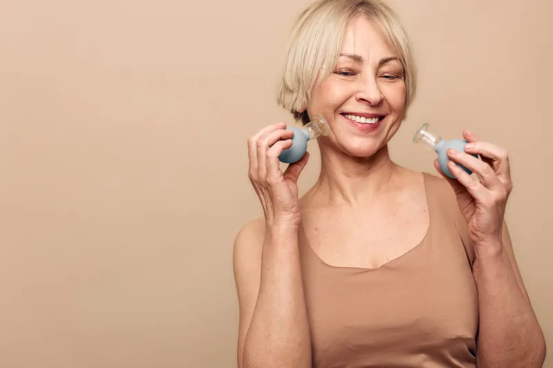 Smiling middle aged woman with short blonde hair holds two facial massage rollers, enjoying skincare routine against beige background. Concept of wellness, self care, and anti aging beauty.