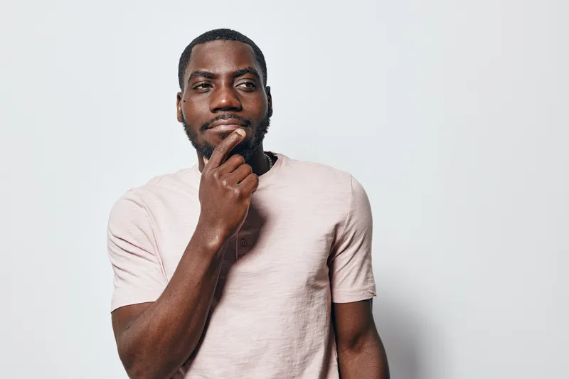 African American man with dark skin and beard wearing light pink t shirt standing thoughtful with hand on chin against white background in studio portrait.