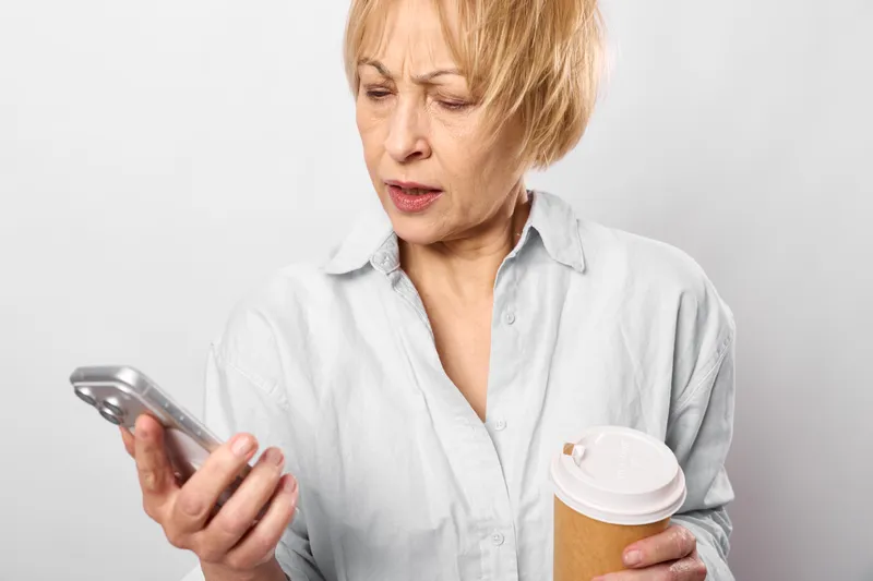 Elderly woman with smartphone and coffee cup, isolated on white background, shows stress and worry while looking at screen, concerned expression, indoor portrait, senior technology