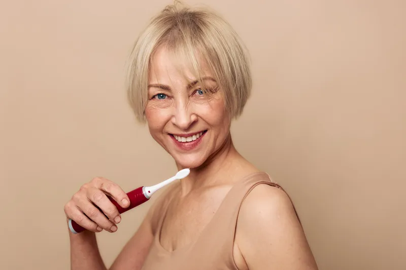 Smiling mature woman with short blonde hair uses electronic toothbrush on beige background. Concept of oral hygiene, dental care, healthy smile, and personal wellness routine.