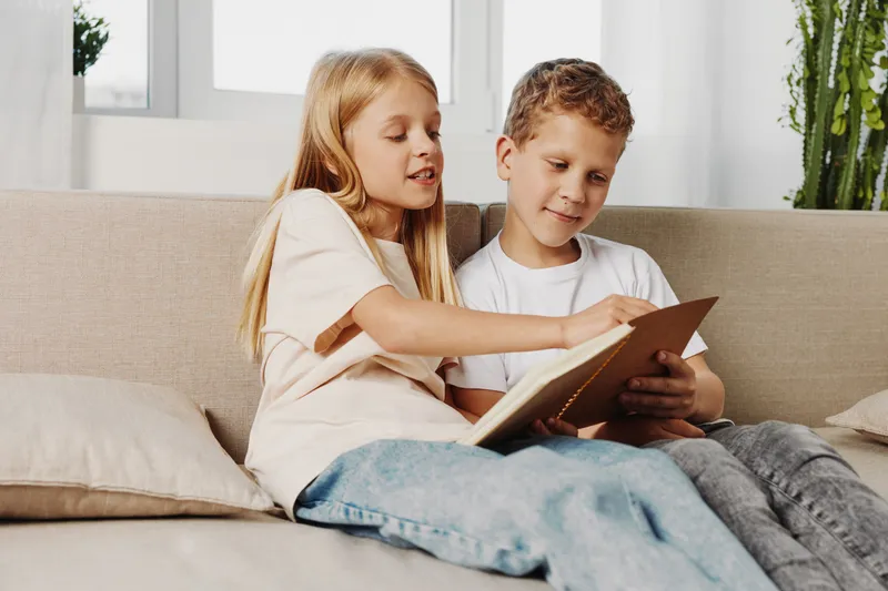 Two children sitting on a couch reading a book together in a bright living room. The girl points at the pages while the boy attentively listens. Concept of learning, childhood, and family time.