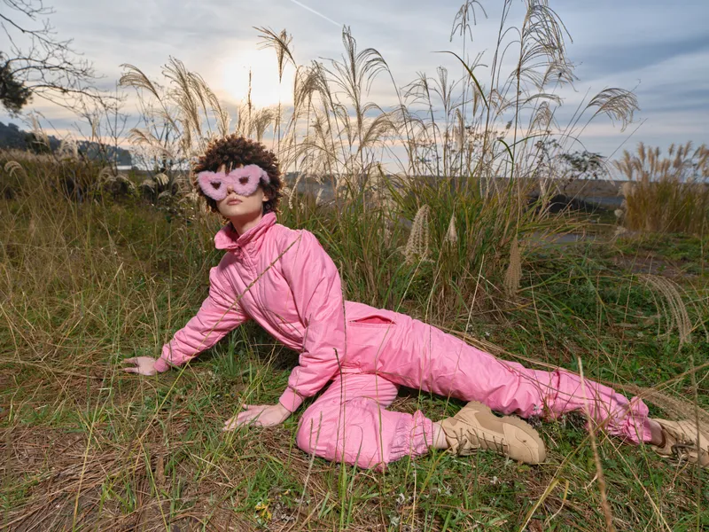 Person in pink jumpsuit and mask lying on a grassy field at sunset, playful outdoor cosplay look with dramatic pose and natural background, rich color and texture