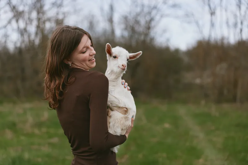 Smiling woman in a brown top holds a goat kid outdoors, sunlight warms the field, capturing a tender rural moment of lifestyle, animal companionship, and wellbeing in a natural setting.