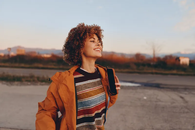 Smiling woman lifestyle outdoors near river at sunset, curly hair, orange jacket, casual travel walk, warm golden daylight, striped sweater, soft blurred landscape, calm evening mood