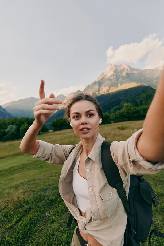 A woman hikes outdoors with a backpack in a mountain landscape, taking a selfie. Bright natural light conveys an active, outdoor lifestyle and a sense of adventure.