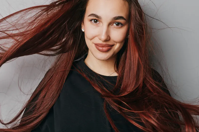 Woman with long red hair smiling and looking at camera with natural makeup and black top on white background. Portrait of happy female with flowing hair and earrings.