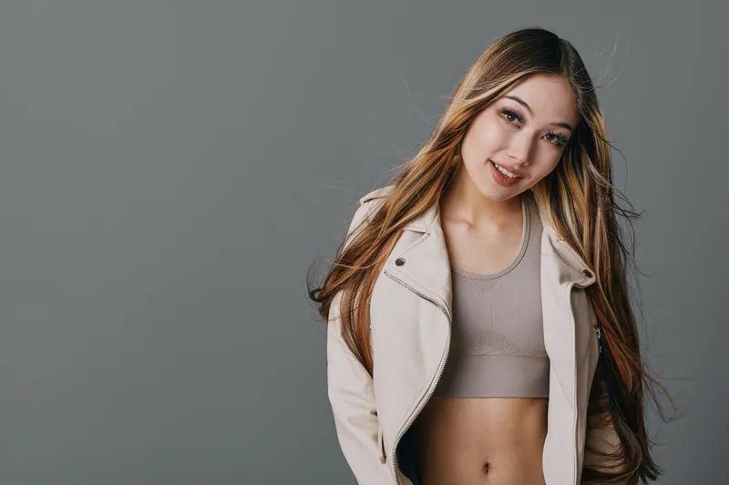 Young woman with long hair smiling wearing a beige jacket and crop top against grey background posing casually with natural makeup and relaxed expression in studio photography.
