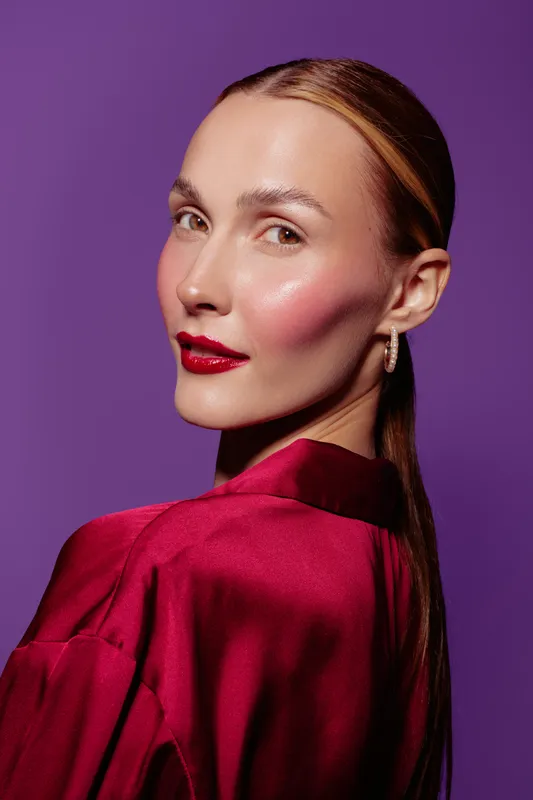 Portrait of a confident woman with glossy red lips and smooth skin wearing a red satin blouse against purple background. Concept of beauty, fashion, and elegance.