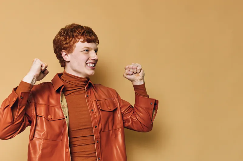 Young man with red hair smiling and raising fists, wearing a brown leather jacket and turtleneck against beige background in studio, expressing excitement and joy.