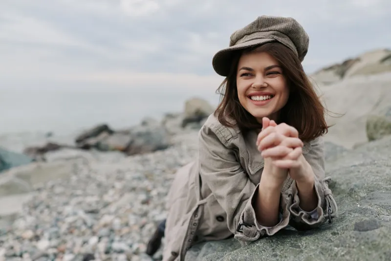 Smiling young woman in casual outfit and hat relaxing on rocky beach. She looks happy and carefree with natural background of stones and cloudy sky. Concept of outdoor leisure and joy.