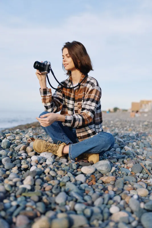 Photographer sits on a pebble beach with camera in hand, wearing a plaid shirt and jeans, capturing seaside scenery with relaxed focus under a clear blue sky.