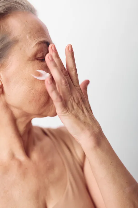 Mature woman applies face cream on cheek with fingers in bright studio. Skincare routine, healthy skin care, anti aging concept with natural beauty and self care for older adults.