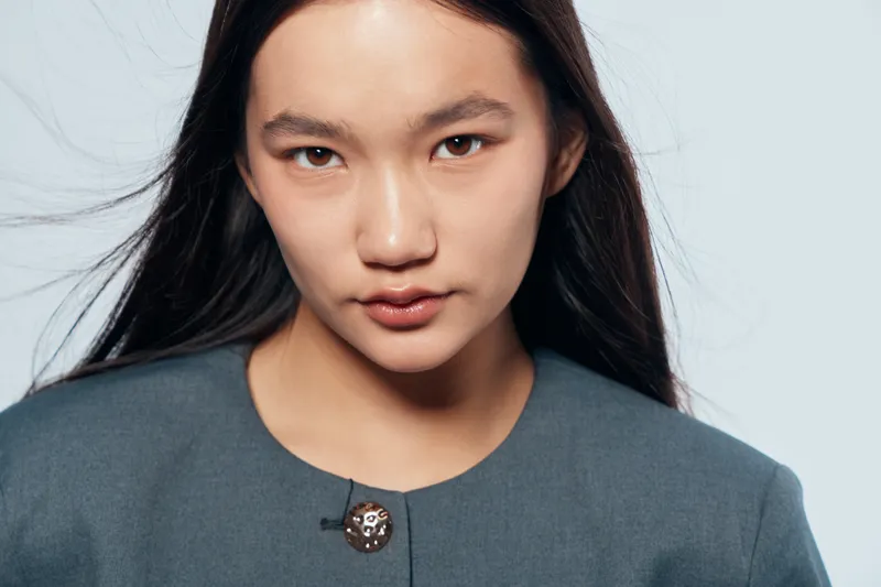 Young Asian woman with long dark hair wearing blue top standing against light background looking seriously at camera with natural makeup and confident expression in studio portrait.